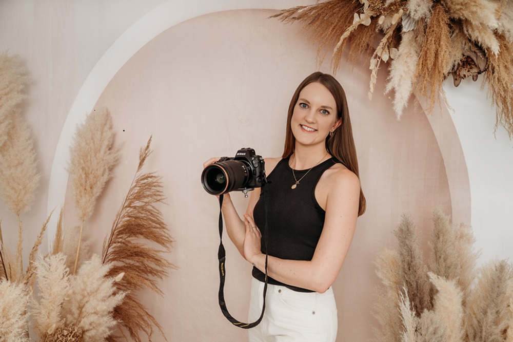 Zu sehen ist die Fotografin von Piuma Photography. Sie steht lächelnd mit einer Kamera in der Hand vor einem beigen Hintergrund.